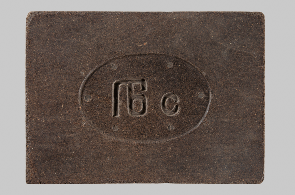 A solid brick of compressed tea, dark brown in colour, with a large monogram impressed in the centre with the Russian characters 'ПБ с'. Photographed against a grey background.